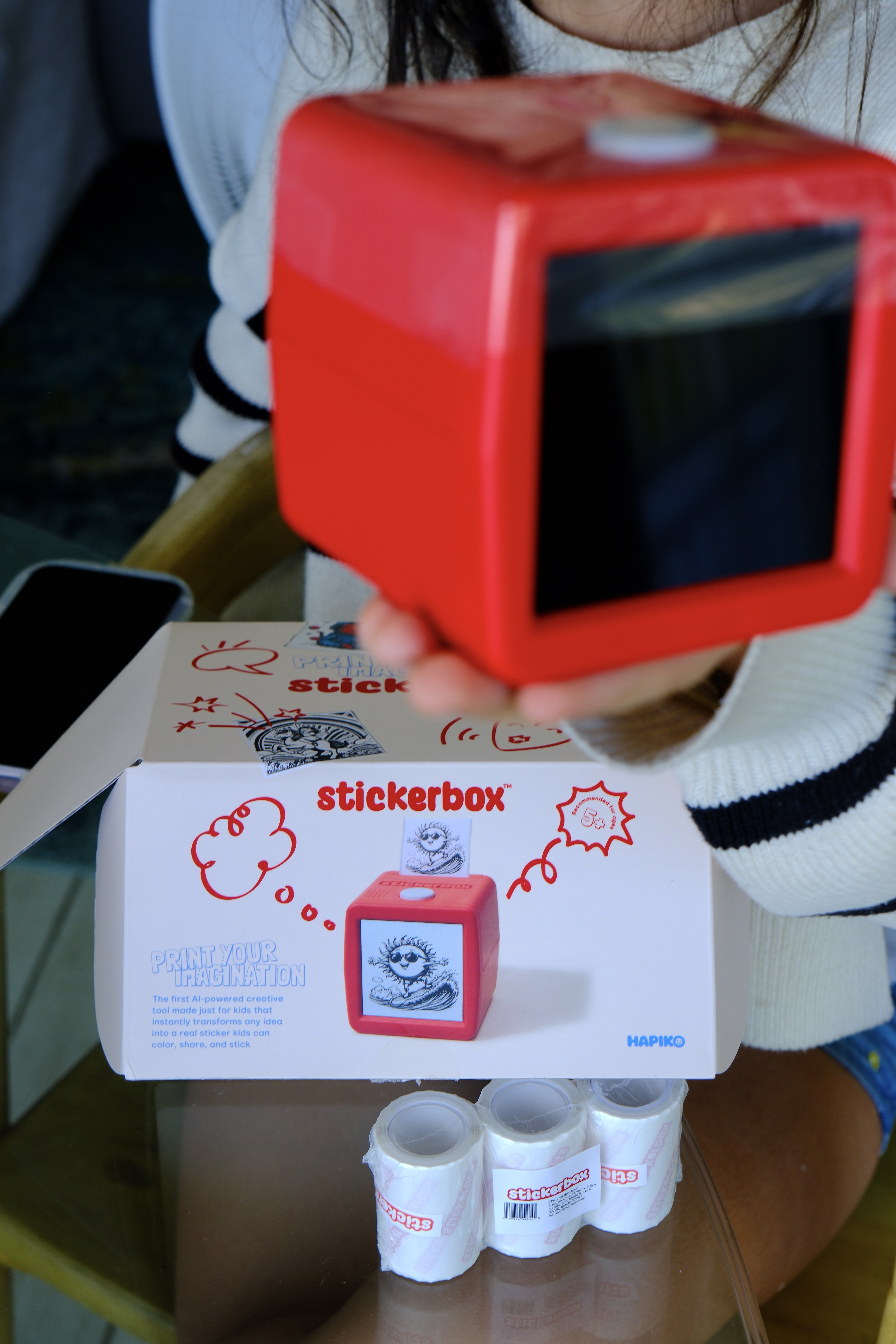 Red Stickerbox AI sticker printer held up above its box and paper refill rolls. Stickerbox promo code SALTYVAGABONDS10 for $10 off at stickerbox.com